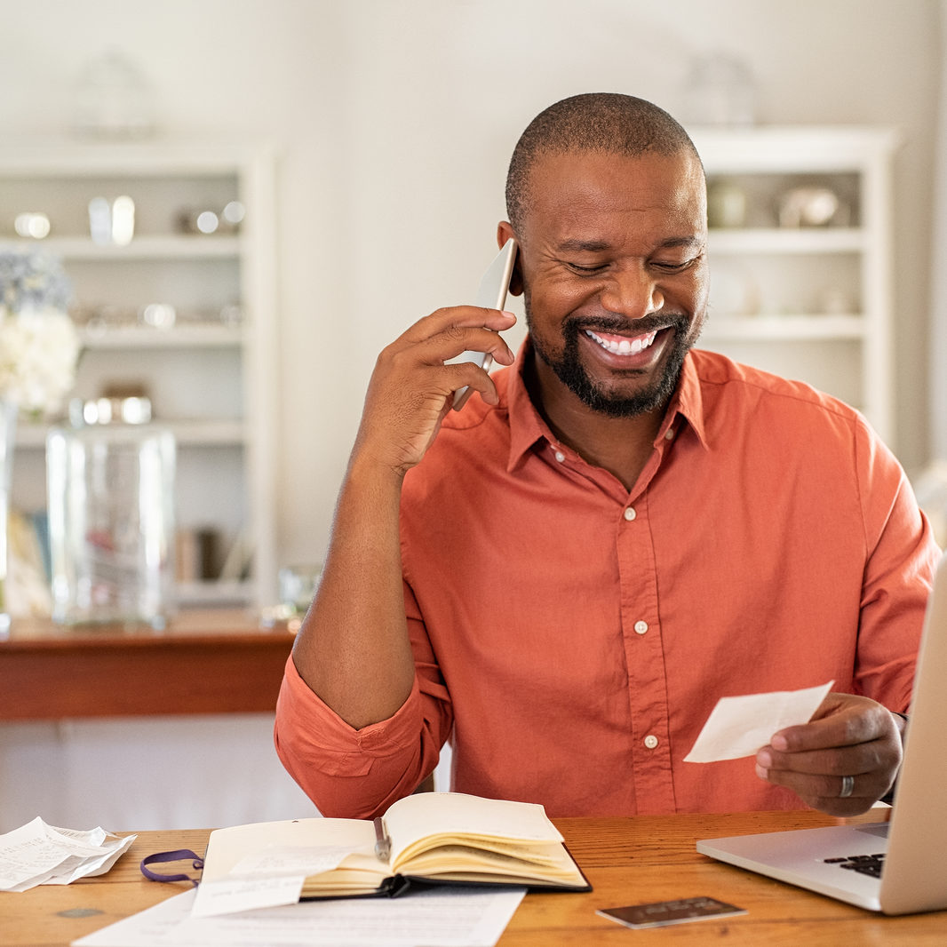 Smiling african man using laptop at home while checking home finance. Happy mature man looking at invoice while talking on phone with bank. Man checking receipt and bill while talking at phone.